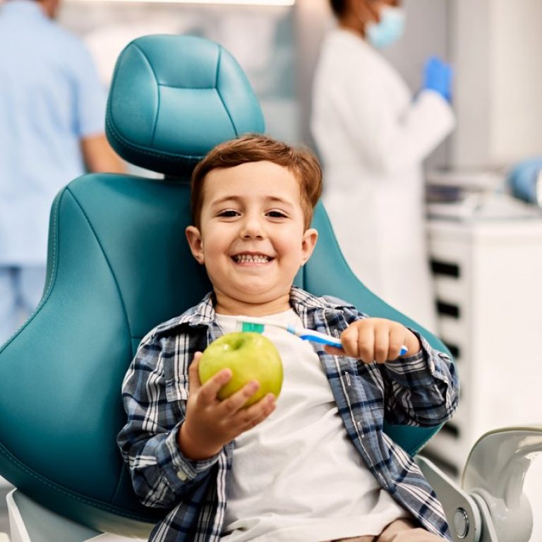 Happy kid learning to brush teeth while using toothbrush and apple at dentist's office and looking at camera.